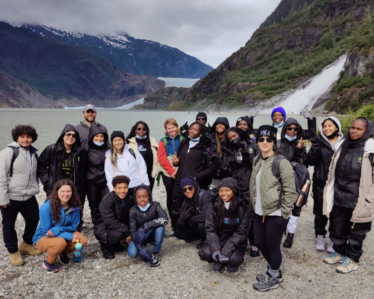 A group of highschool students posing for a photo in front of a lake. A glacier and waterfall can be seen in the background