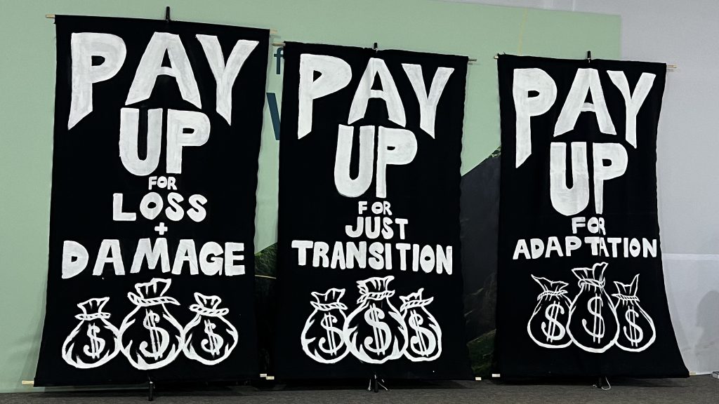 A photo of signs that read "Pay up for loss + damage," "Pay up for just transition," and "Pay up for adaptation" 